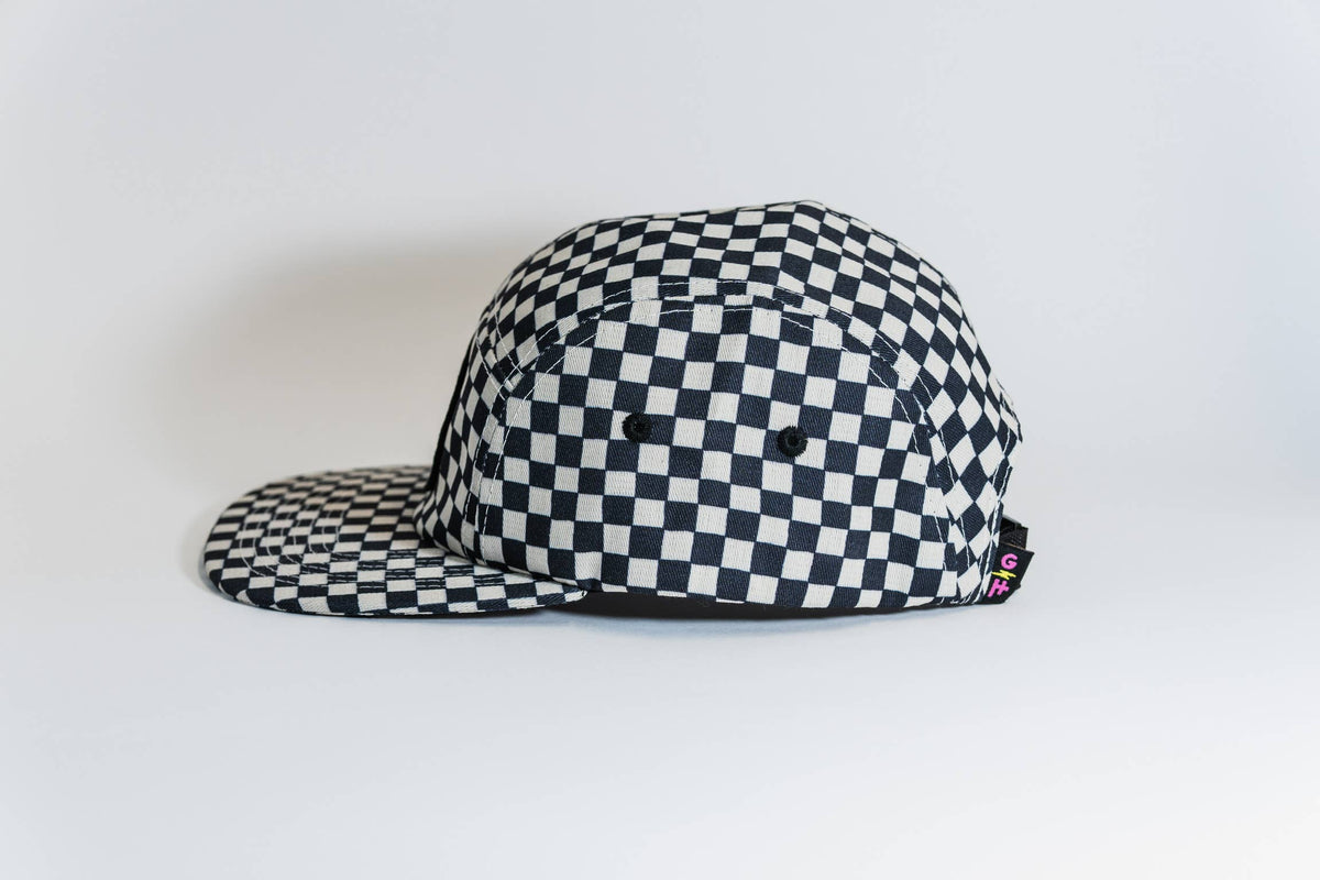 The Steezy Checkered 5-Panel Hat is shown in black and white with a flat brim, repeating square pattern, and a small black tag with colored letters; designed by a women-owned small business.