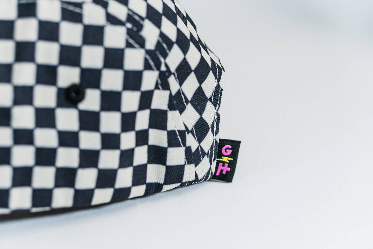 The Steezy Checkered 5-Panel Hat is a black and white checkered outdoor cap with a small black tag showing a pink G, yellow I, and green F. Designed by a women-owned small business, it pops against a plain white background.