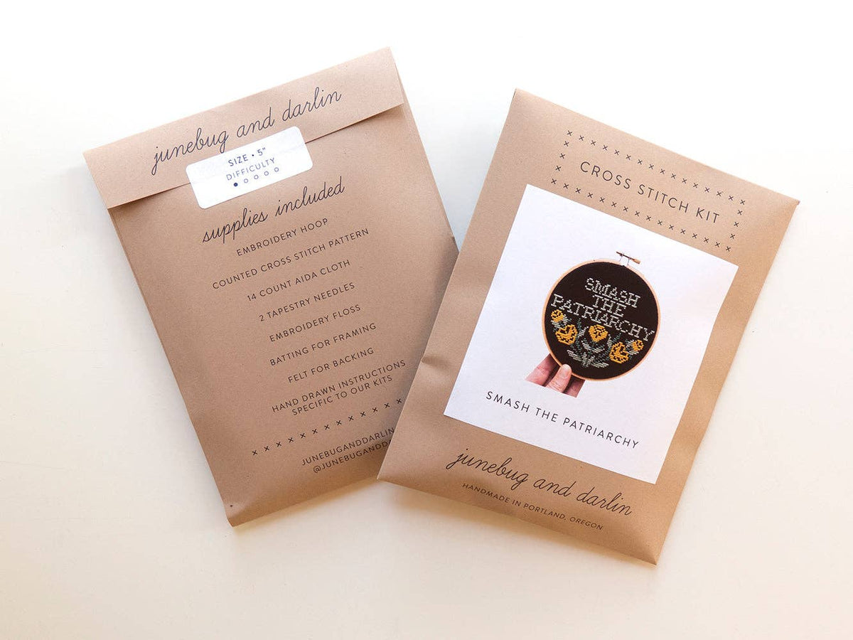Two brown paper envelopes for the beginner-friendly Smash the Patriarchy Cross Stitch Kit; one shows included supplies, the other displays a completed hoop reading "Smash the Patriarchy" with floral accents.