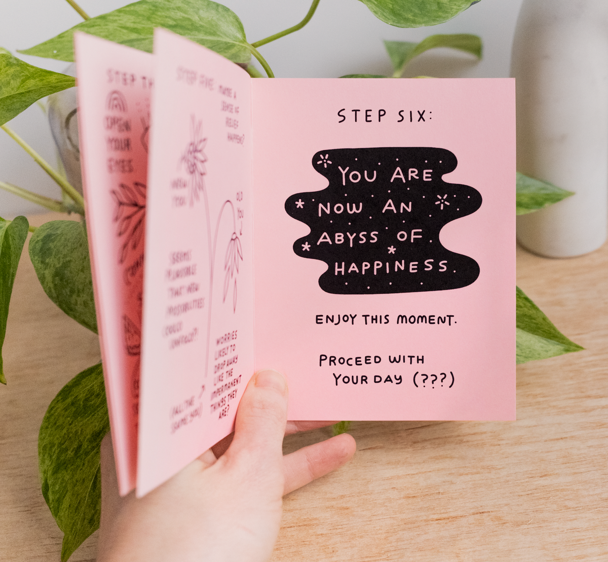A hand holds the Little Happiness Zine open to a page that reads, "STEP SIX: YOU ARE NOW AN ABYSS OF HAPPINESS. Enjoy this moment. Proceed with your day (???)" in decorative text, next to an eco-friendly green plant.