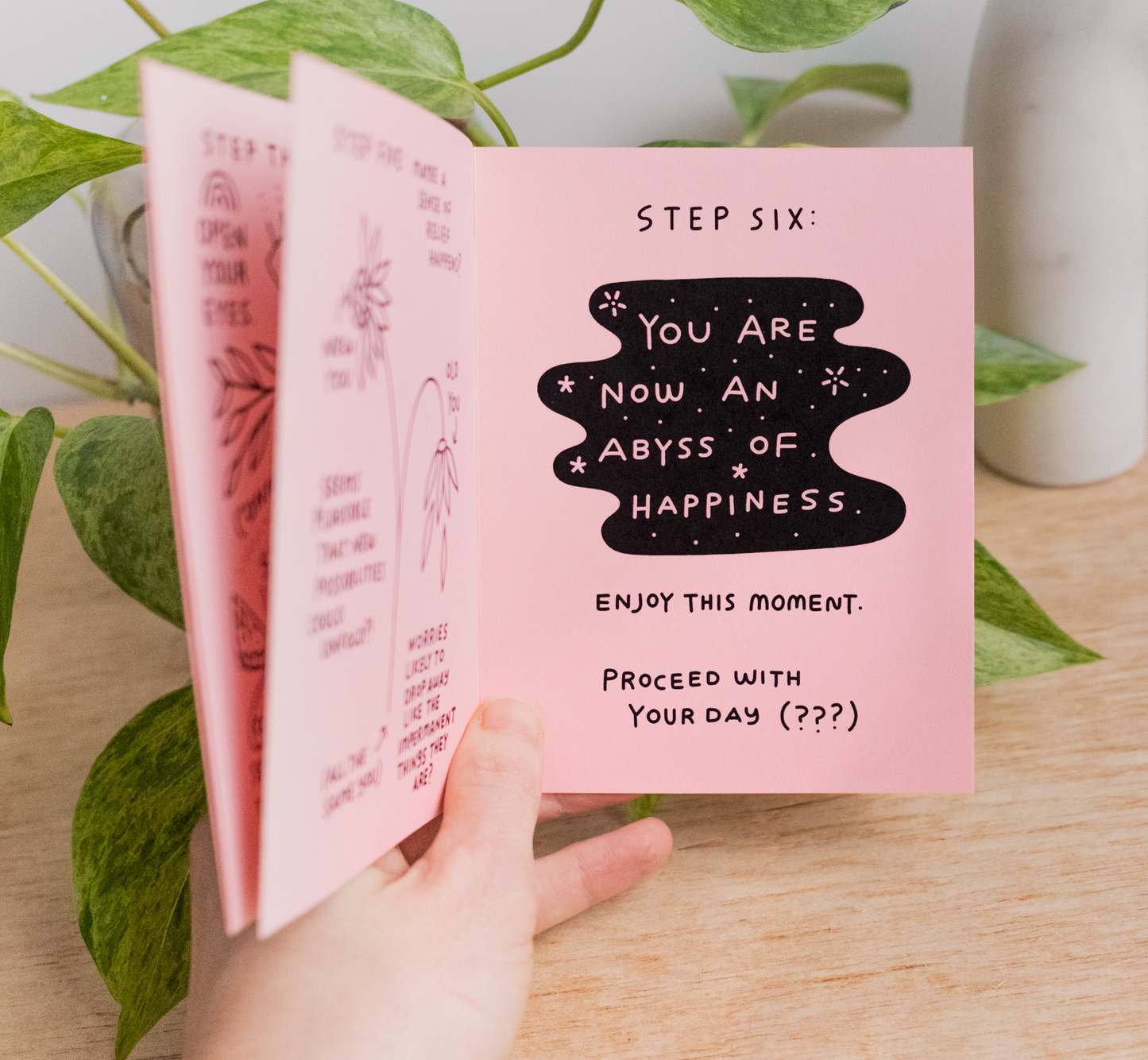 A hand holds the Little Happiness Zine open to a page that reads, "STEP SIX: YOU ARE NOW AN ABYSS OF HAPPINESS. Enjoy this moment. Proceed with your day (???)" in decorative text, next to an eco-friendly green plant.
