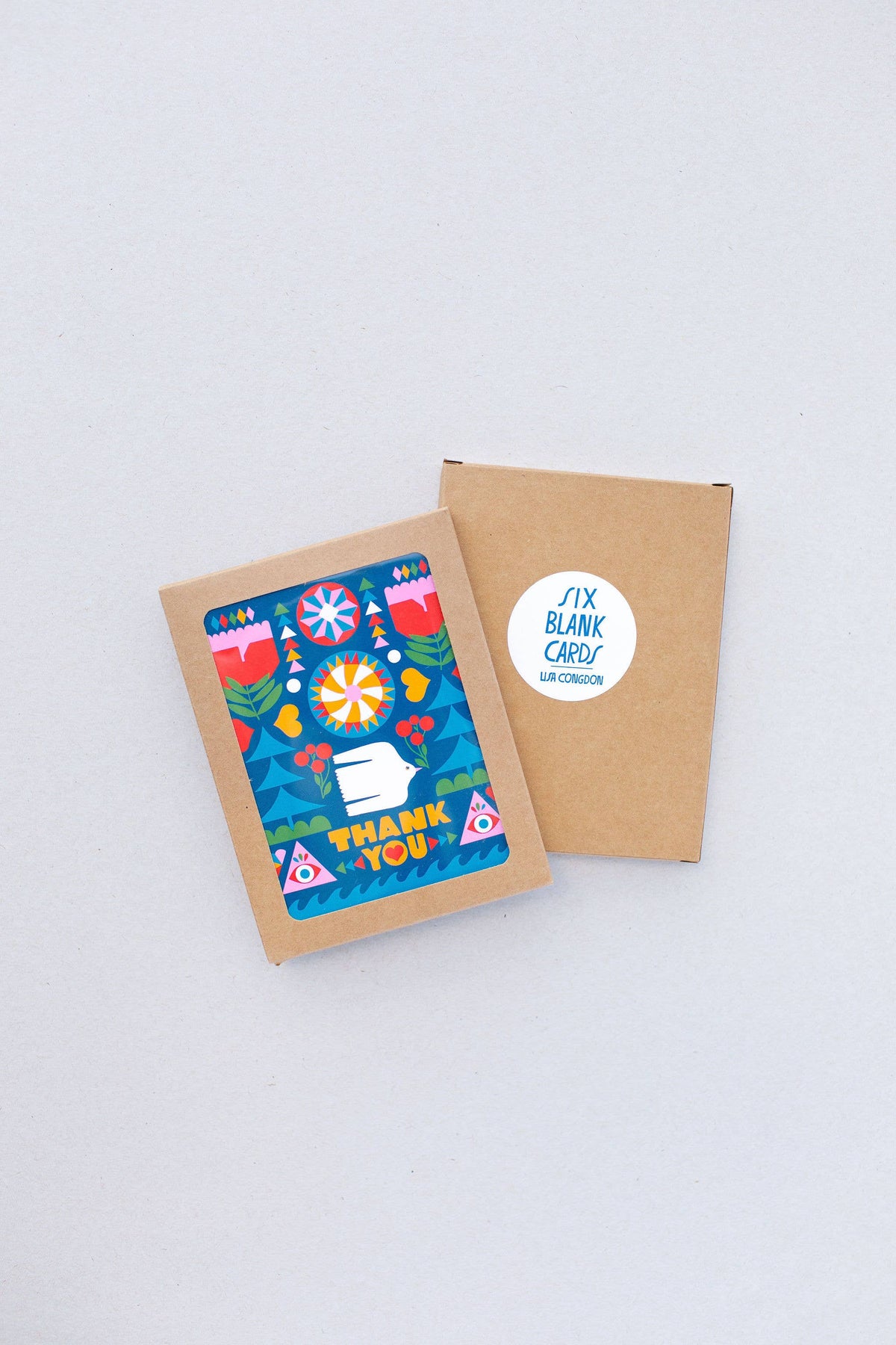 The Folk Flowers Thank You Greeting Card, featuring colorful folk art and geometric designs with "Thank You" text, is partly in a brown envelope. Another brown envelope with a Six Blank Cards sticker lies nearby on a light surface.