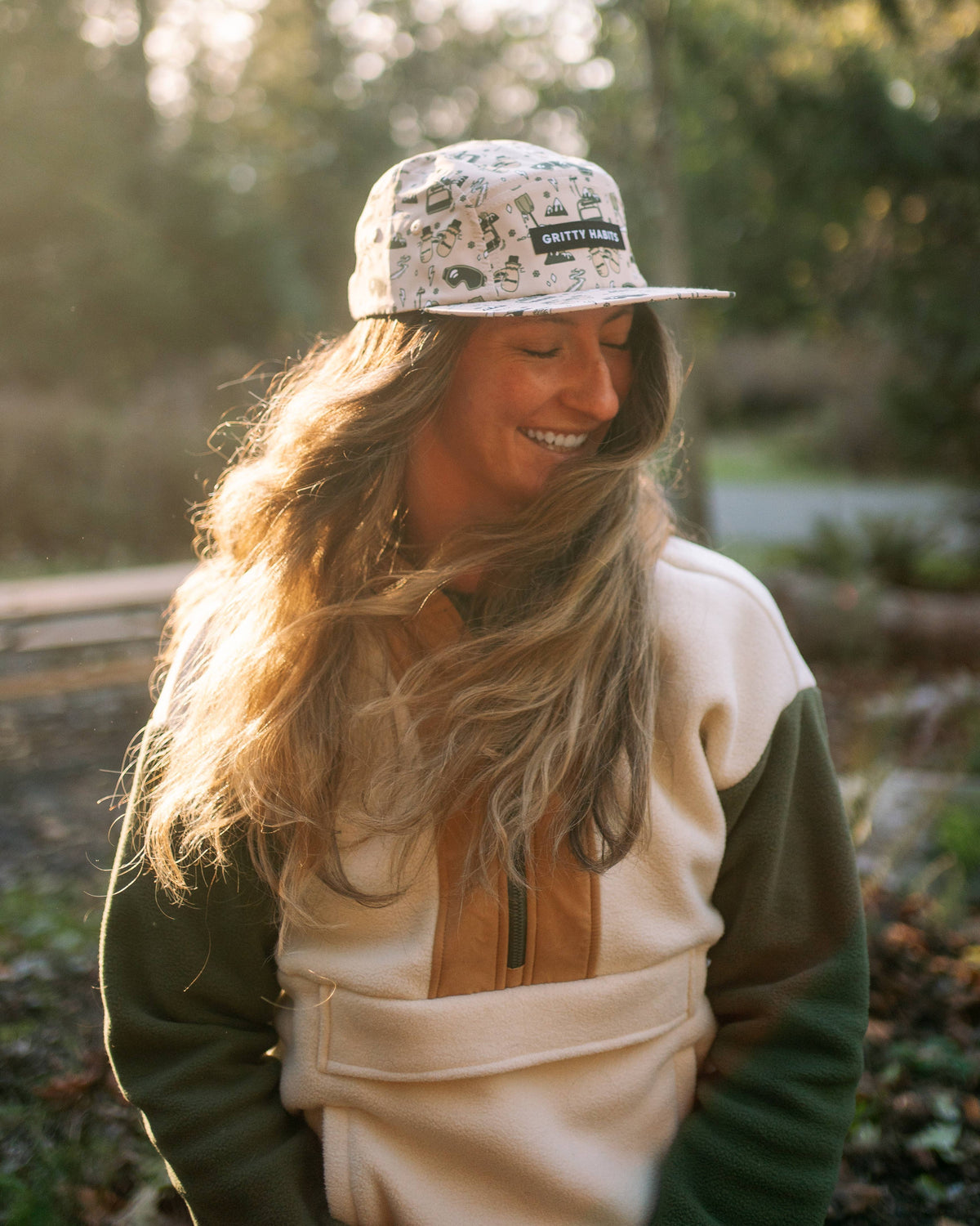 A woman with long, wavy hair smiles outdoors in soft sunlight, wearing a green and cream fleece jacket and the First Chair 5-Panel Hat. Trees and a blurred background highlight the natural setting.