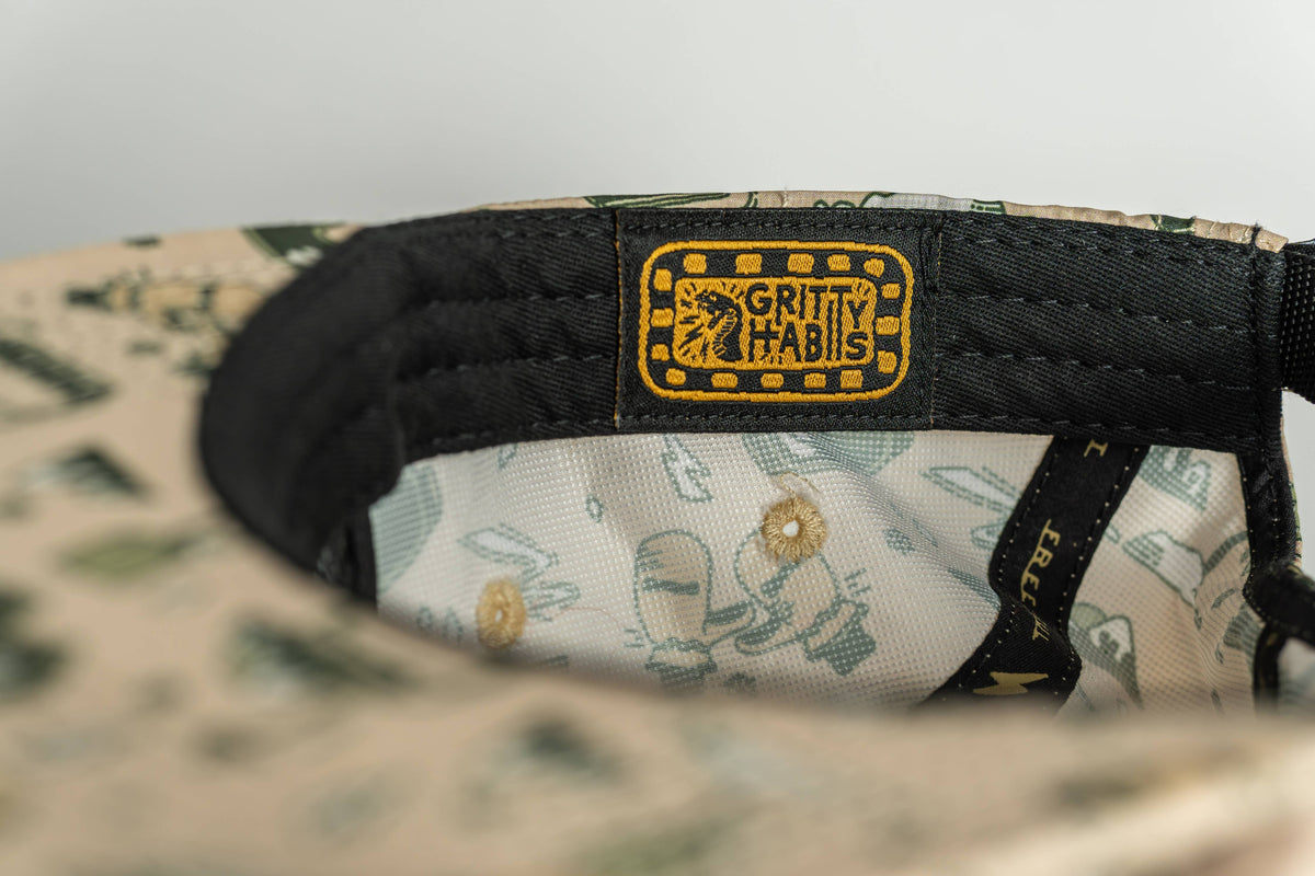 Close-up of the beige First Chair 5-Panel Hat's interior, highlighting green patterns, a stitched yellow and black Gritty Habits label on the black inner band, and patterned performance polyester lining.