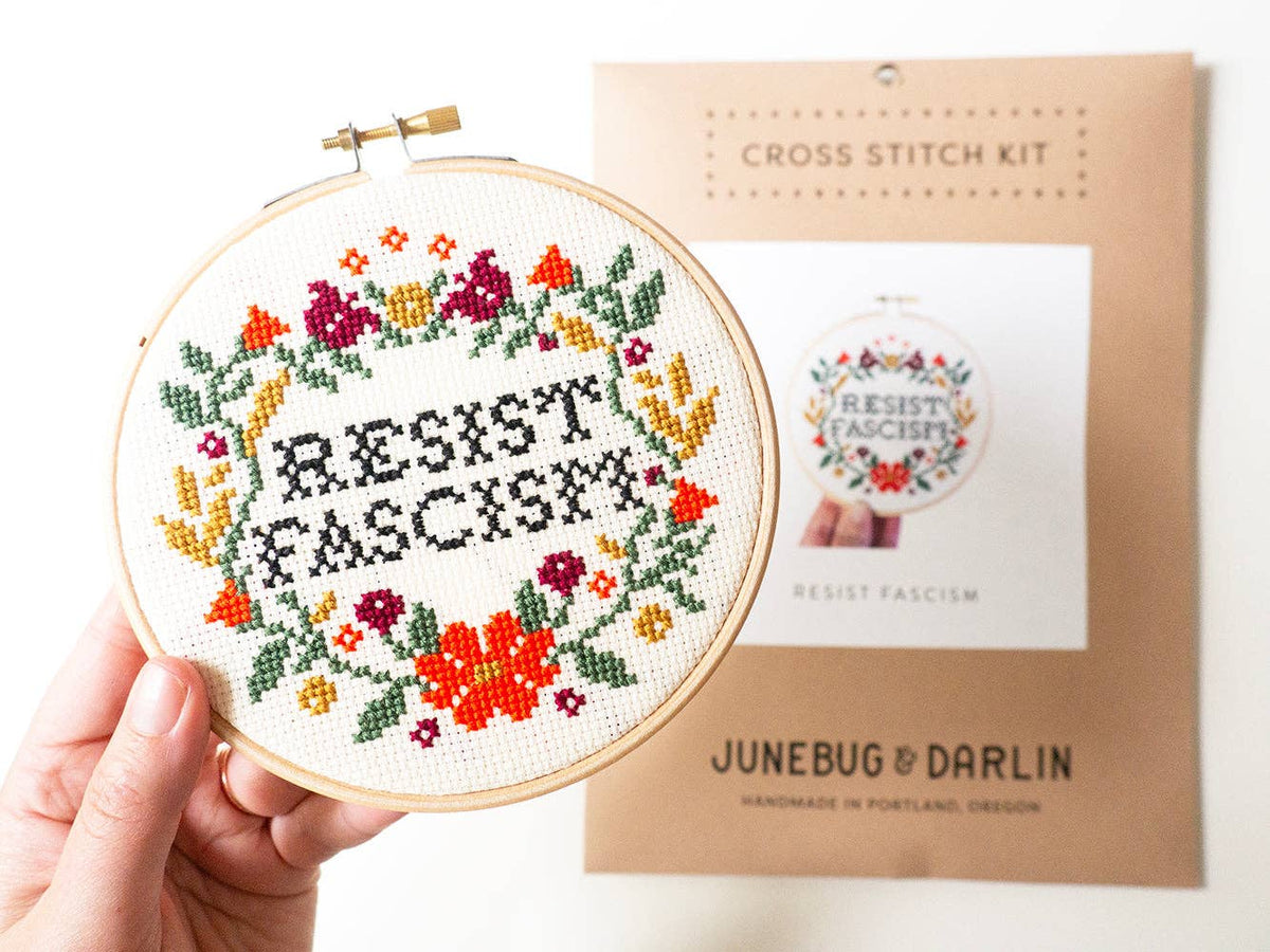 A hand holds the Resist Fascism Cross Stitch Kit, featuring “RESIST FASCIST” stitched in black with colorful florals. The beginner-friendly package behind it shows the same handmade in Portland design.