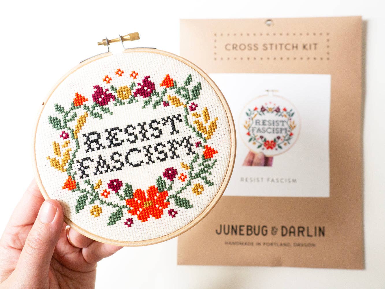 A hand holds the Resist Fascism Cross Stitch Kit, featuring “RESIST FASCIST” stitched in black with colorful florals. The beginner-friendly package behind it shows the same handmade in Portland design.