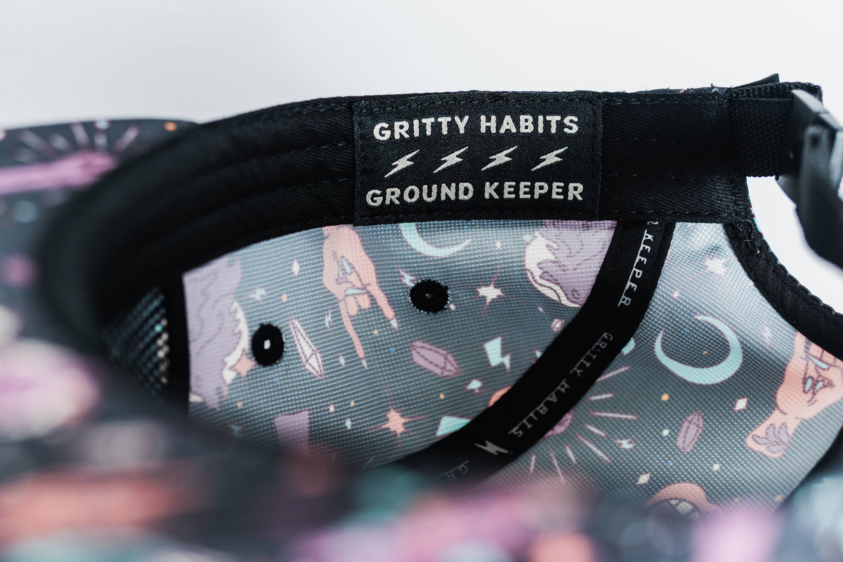 Close-up of the inside of the Gritty Habits x Ground Keeper: Howl Collection 5-Panel Hat, featuring a colorful pattern with moons, stars, and hands. A black label reads "GRITTY HABITS ⚡⚡⚡ GROUND KEEPER." Proudly women-owned.