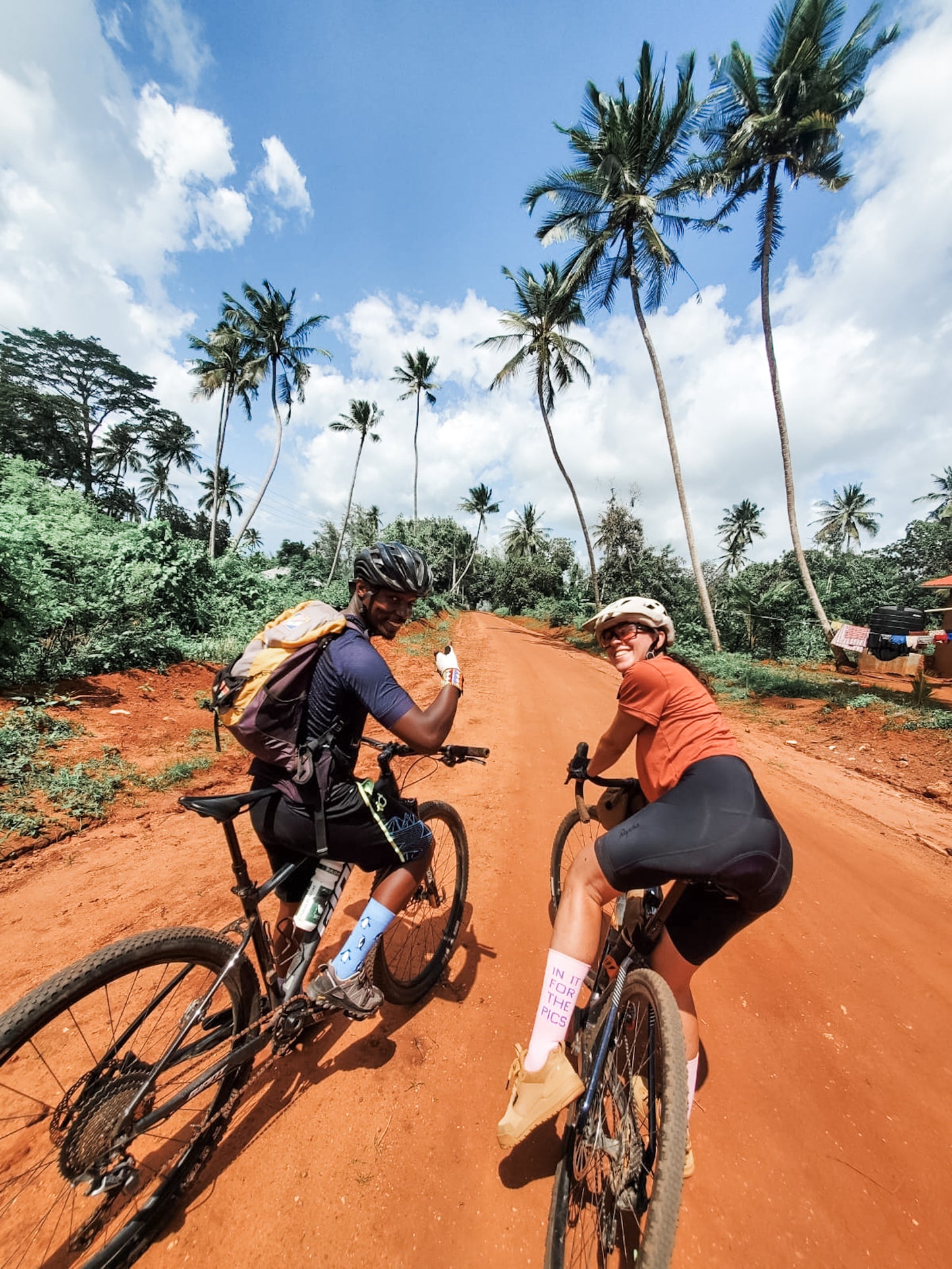 Enjoy the Cycling Safari as two helmeted cyclists smile along a red dirt road lined with tall palm trees beneath a blue sky, surrounded by lush greenery—one pauses to wave at the camera, inviting you to join the adventure.