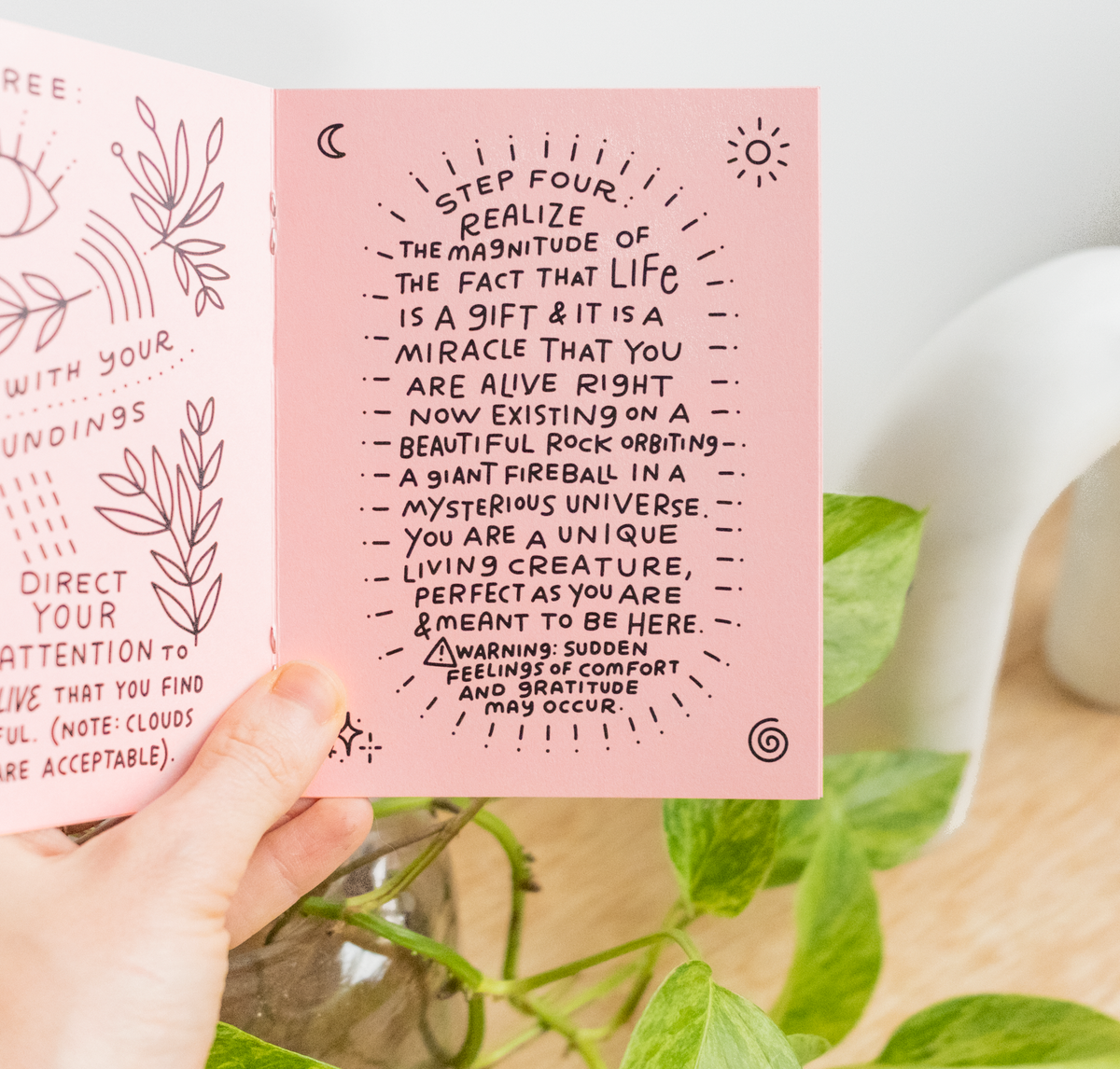 A hand holds open the eco-friendly Little Happiness Zine, featuring handwritten inspirational text and small sun and star doodles. A green plant is in the background, setting a calming vibe.
