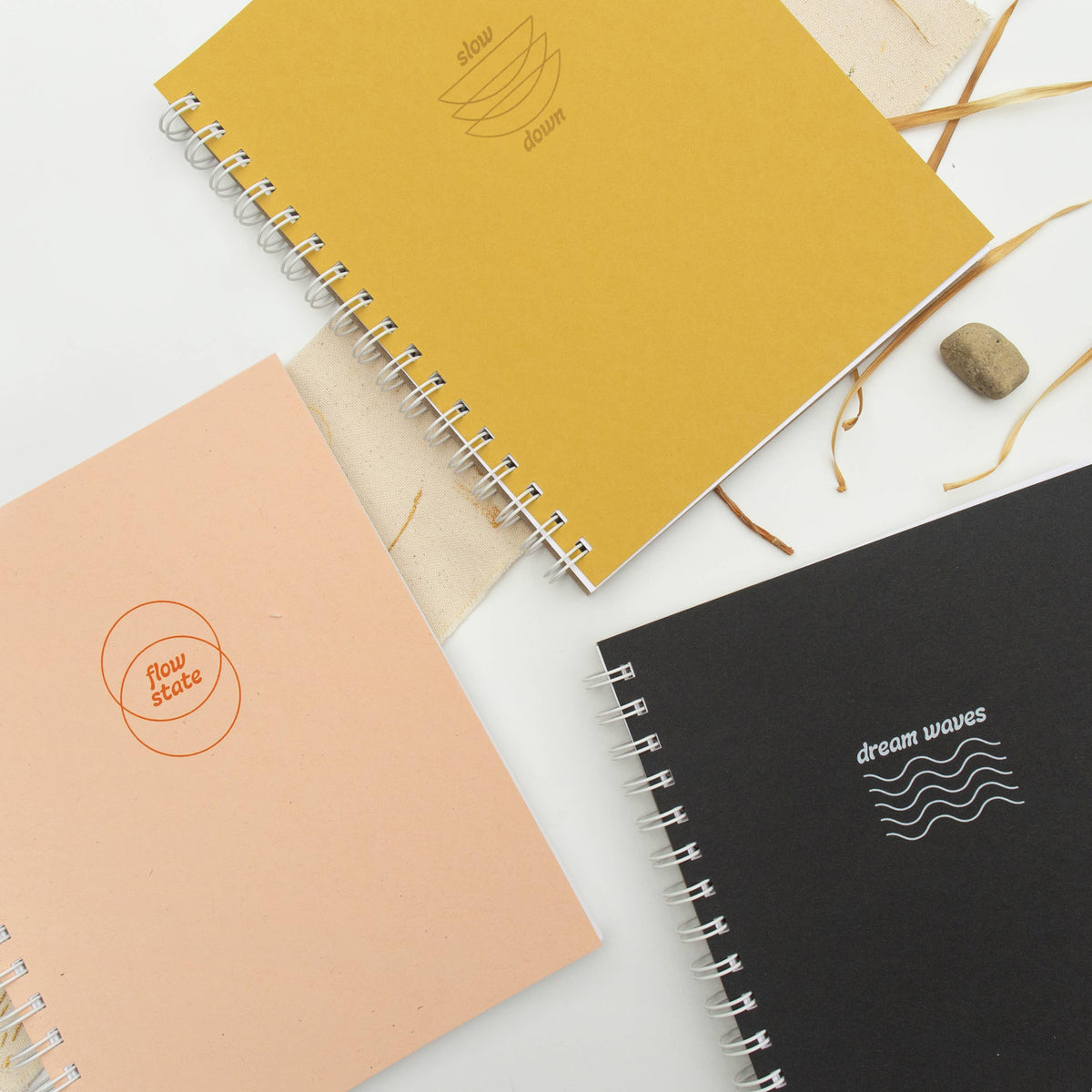 Three spiral-bound notebooks with minimalist covers—one yellow reading slow down, one pink Flow State Guided Journal, and one black dream waves—are displayed on a white surface with natural accents, perfect for sparking creativity.