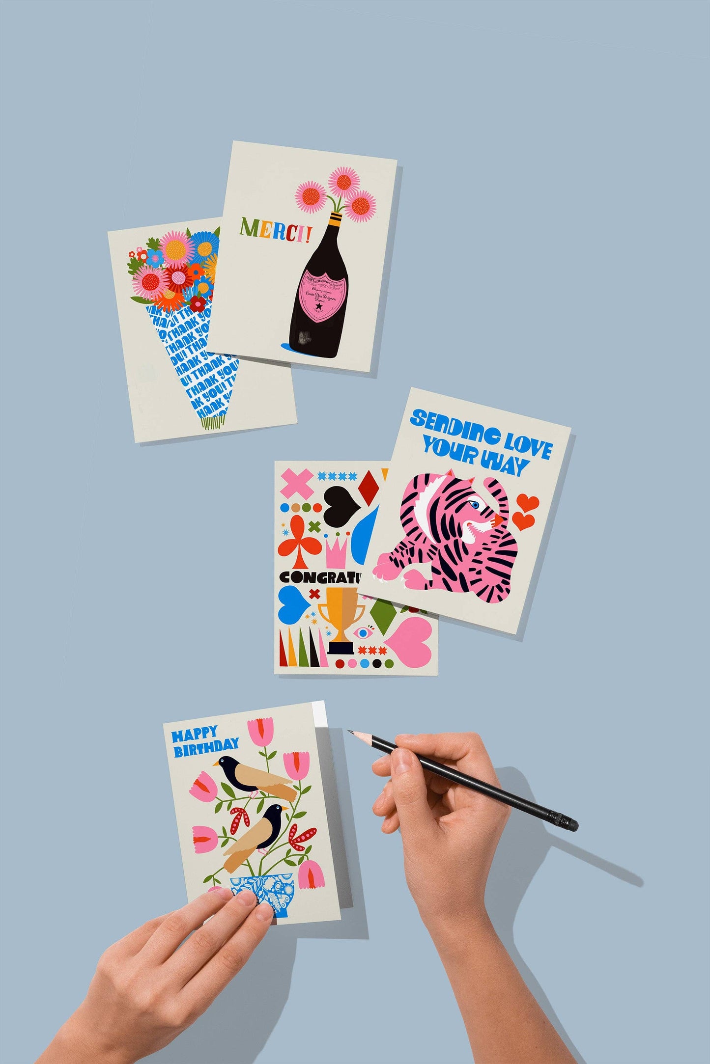 A hand writes on the Sending Love Pink Tiger Greeting Card. Five cards with colorful, playful designs and messages like "Sending Love" are displayed on a light blue background; each card is blank inside.
