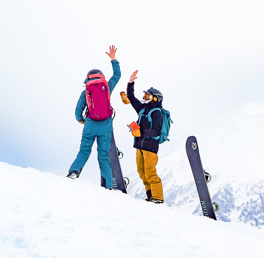 Jen and Slu high-fiving on a snowy ridge in Japan, skis planted beside them