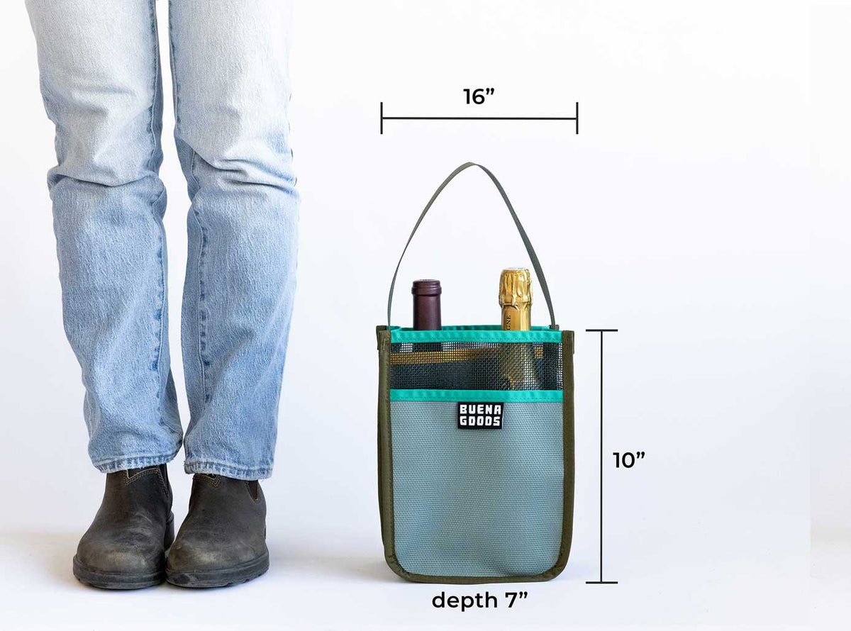 A person stands next to a gray and teal The Two-Bottle-Bag | Cava on the Playa by Buena Goods, which holds two wine bottles and measures 16"W x 10"H x 7"D. The lower legs and boots of the person are visible.