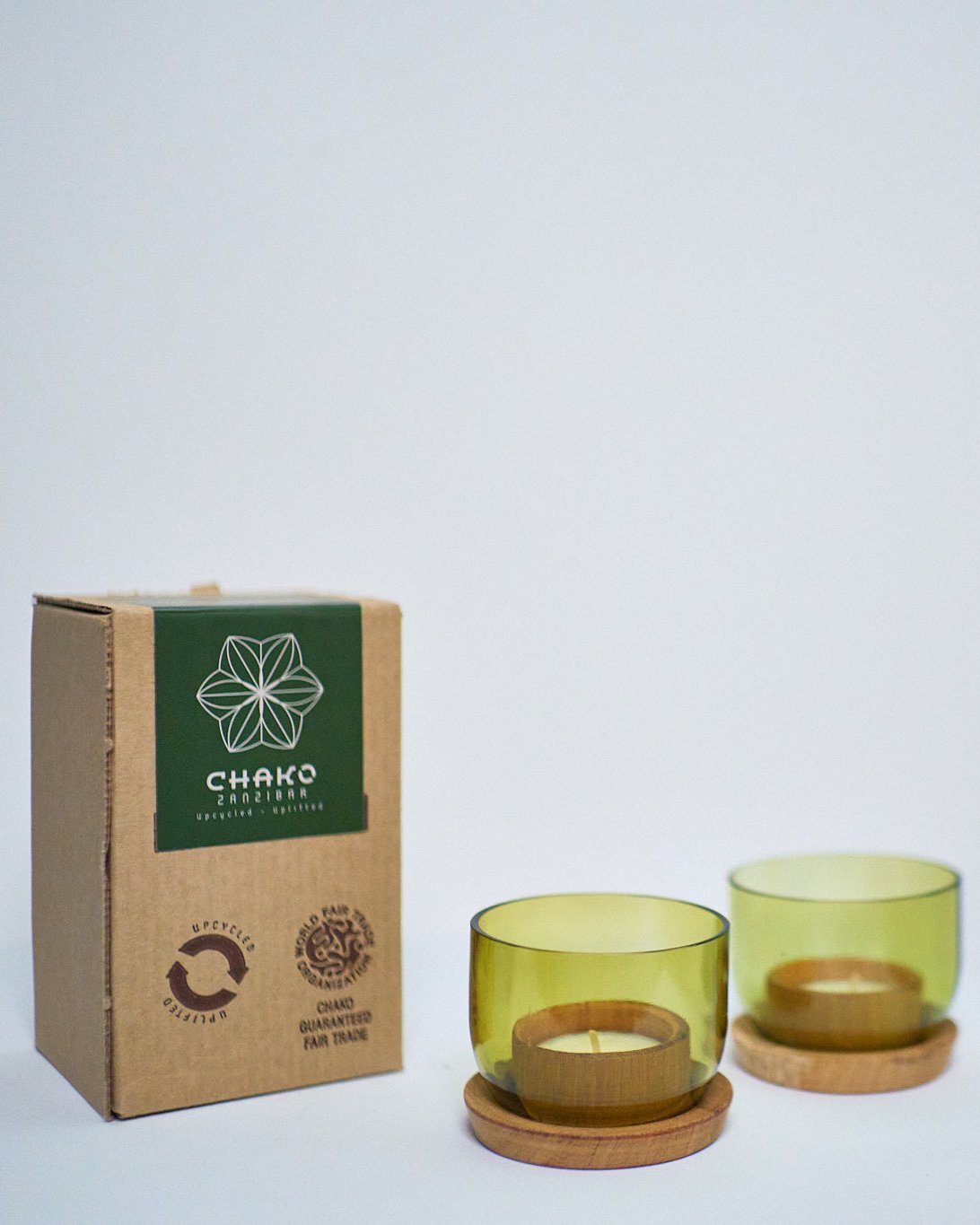 A cardboard box labeled CHAKO stands next to two green-tinted Taa Tea Lights Candle Holders, upcycled from wine bottles with wooden bases and marked with eco-friendly and fair trade decor logos, all on a plain white background.