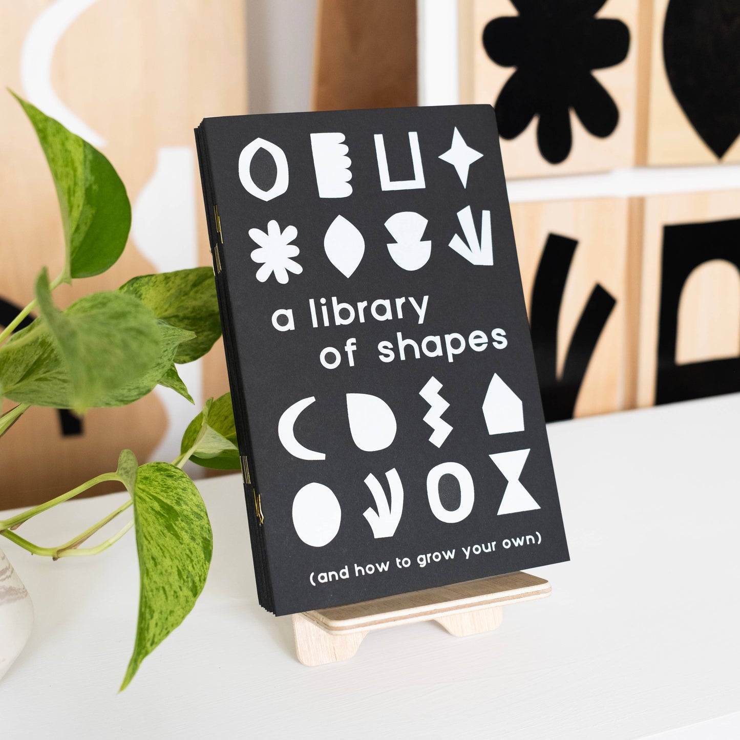 A Library of Shapes Creativity Zine, a black zine with abstract white shapes on the cover, is displayed upright on a small wooden stand beside a green leafy plant.