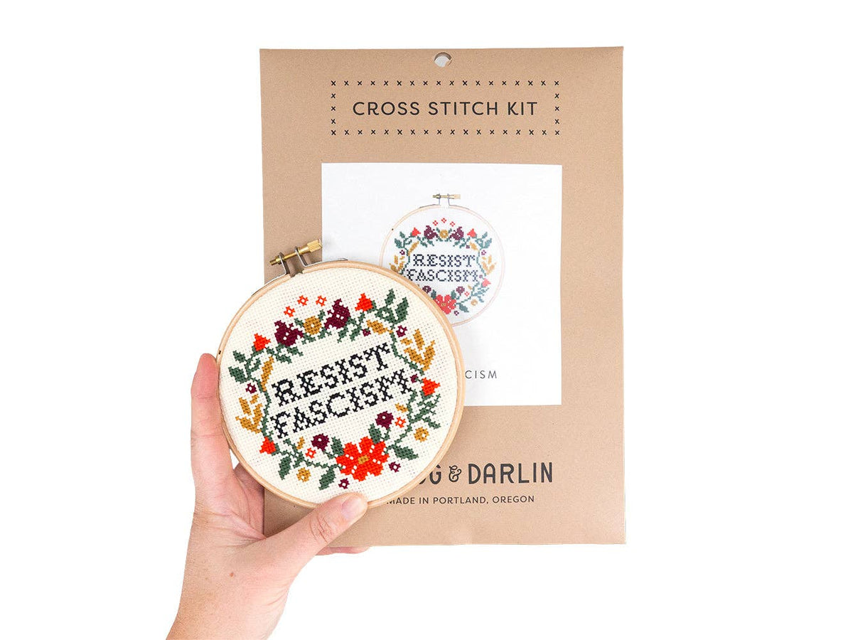 A hand holds a finished Resist Fascism Cross Stitch Kit piece with "RESIST FASCISM" and colorful flowers; the beginner-friendly kit is handmade in Portland, and its packaging can be seen in the background.