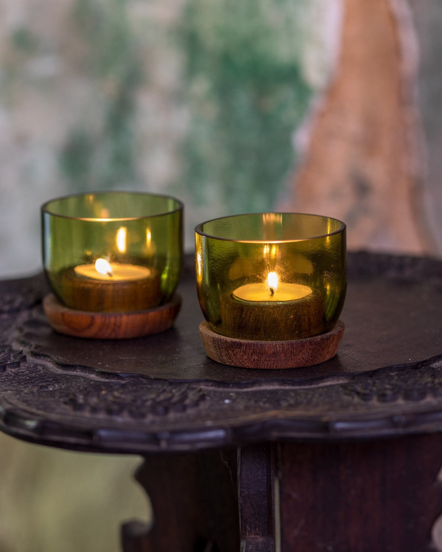 Two Taa Tea Lights Candle Holders, crafted from upcycled green glass with wooden bases, hold small tea light candles burning on an ornate dark wood table against a blurred green and beige backdrop.