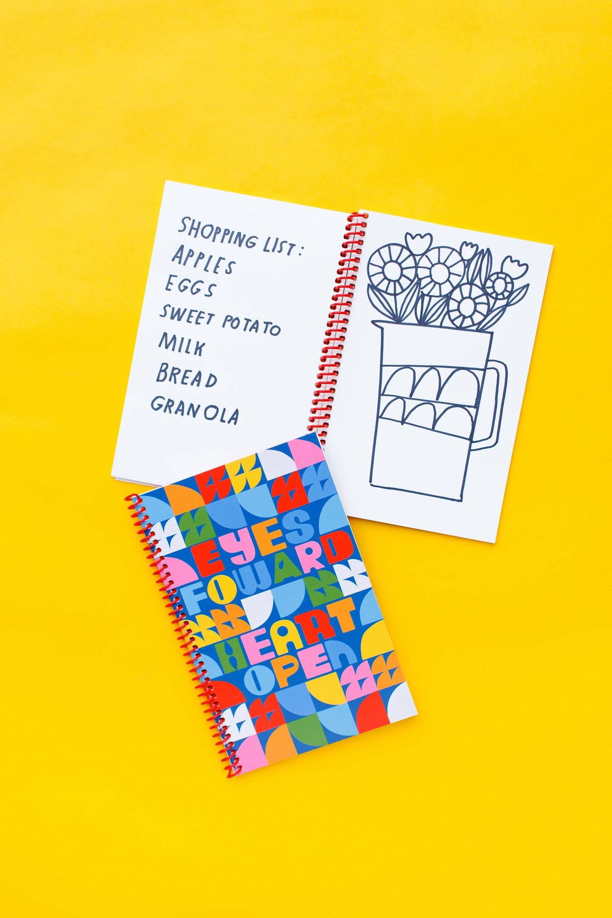 Two spiral journals rest on a yellow surface; one is open with a shopping list and flower vase sketch, while the closed Eyes Forward, Heart Open Journal displays its colorful geometric cover with the phrase “EYES FORWARD, HEART OPEN.”.