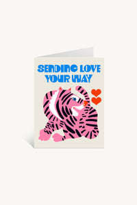 The Sending Love Pink Tiger Greeting Card features Lisa Congdon’s colorful pink and purple tiger with two red hearts and bold blue "SENDING LOVE YOUR WAY" text on a white background; blank inside for your message.