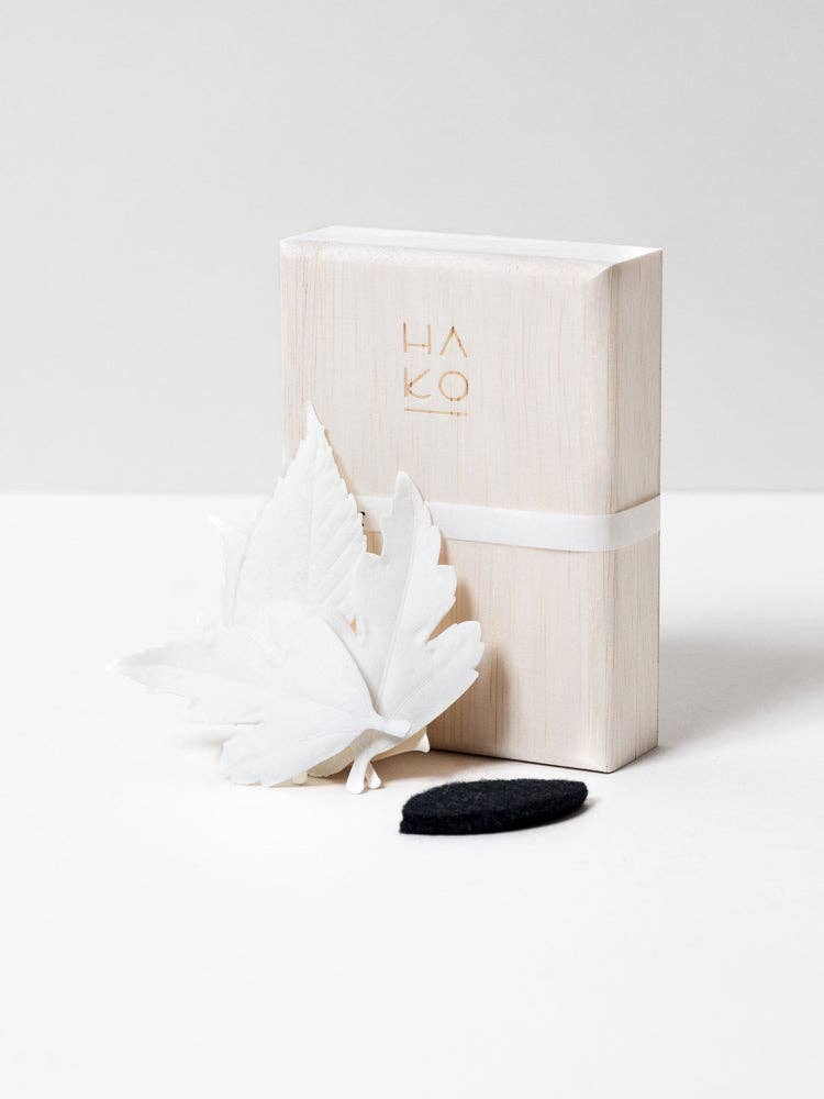 An upright light wooden box engraved with “HAKO” stands behind two white artificial leaves and a small black oval object, evocative of Japanese incense and the refined design of HA KO Paper Incense.