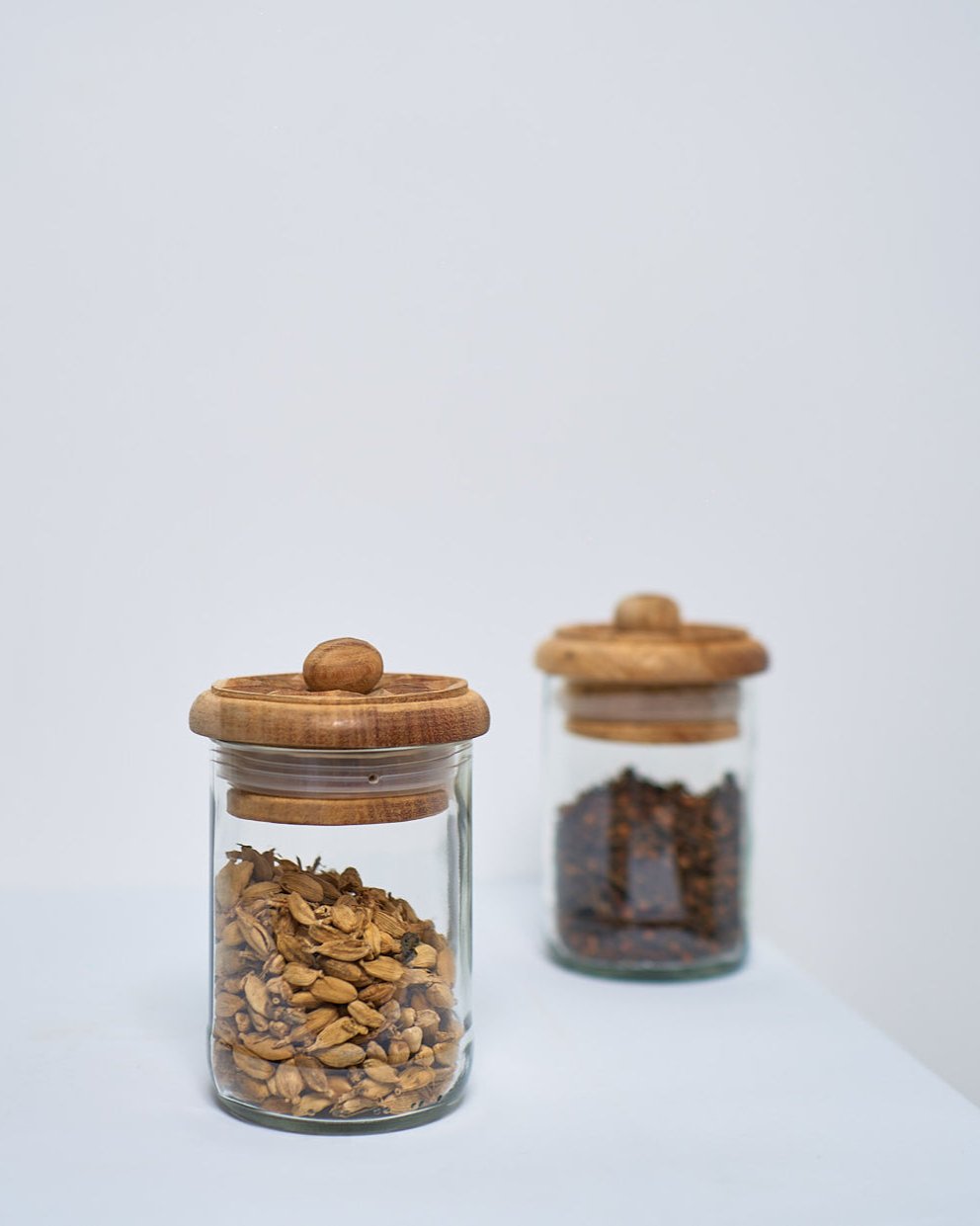 Two X Large Karafuu Upcycled Glass Jars with wooden lids rest on a white surface; one holds brown grains, the other a darker, coarse substance. The background is plain and light.
