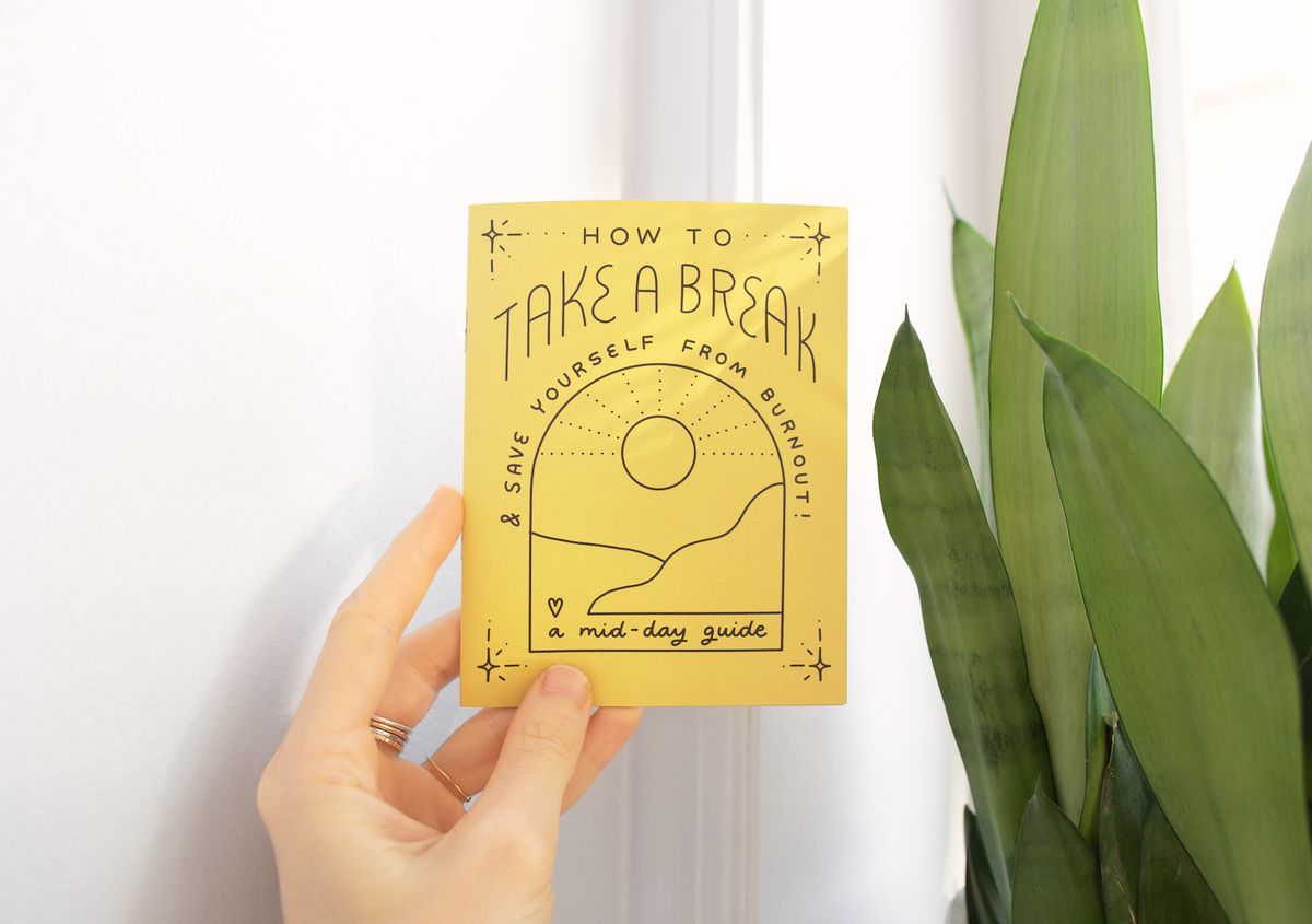 A hand holds the Take a Break Zine, a yellow booklet titled "How to take a break & save yourself from burnout: a mid-day guide," featuring a simple sun and landscape illustration. A green plant appears on the right.