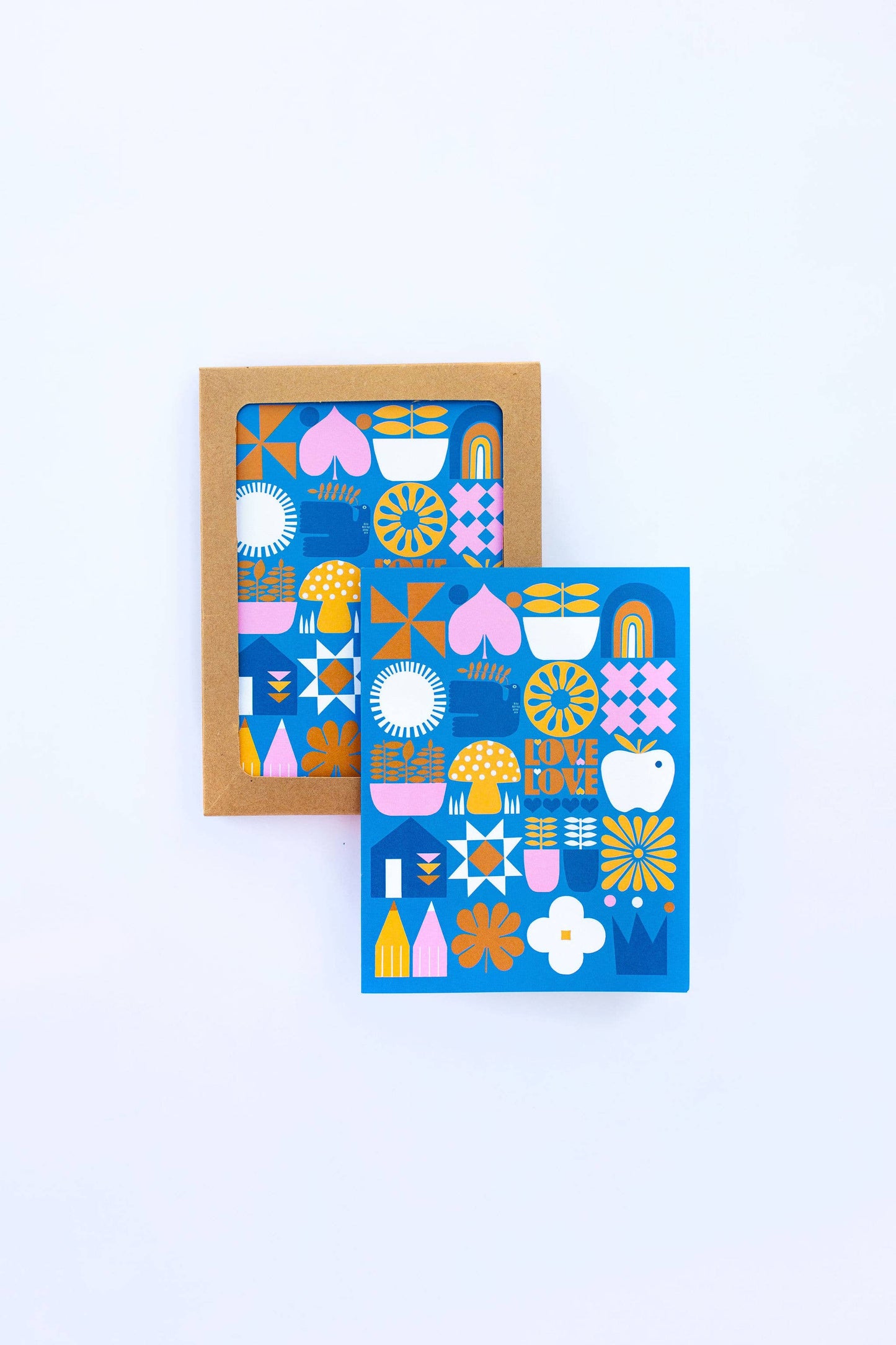 Two colorful Love Love Mushroom Greeting Cards with geometric and floral designs in blue, pink, yellow, and white are on a white background; one blank card is shown inside a brown envelope.