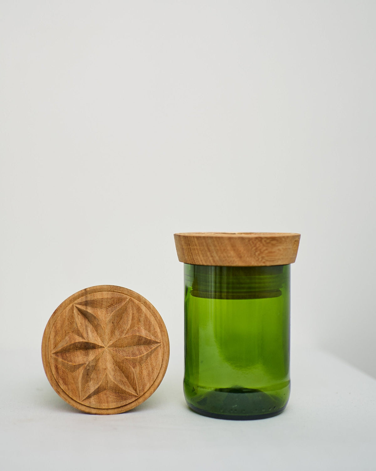 The Hiliki Upcycled Glass Jars | Large Set features a green glass jar with a carved geometric wooden lid. The fair trade lid is shown on top of the jar and propped upright beside it, all displayed on a white surface.