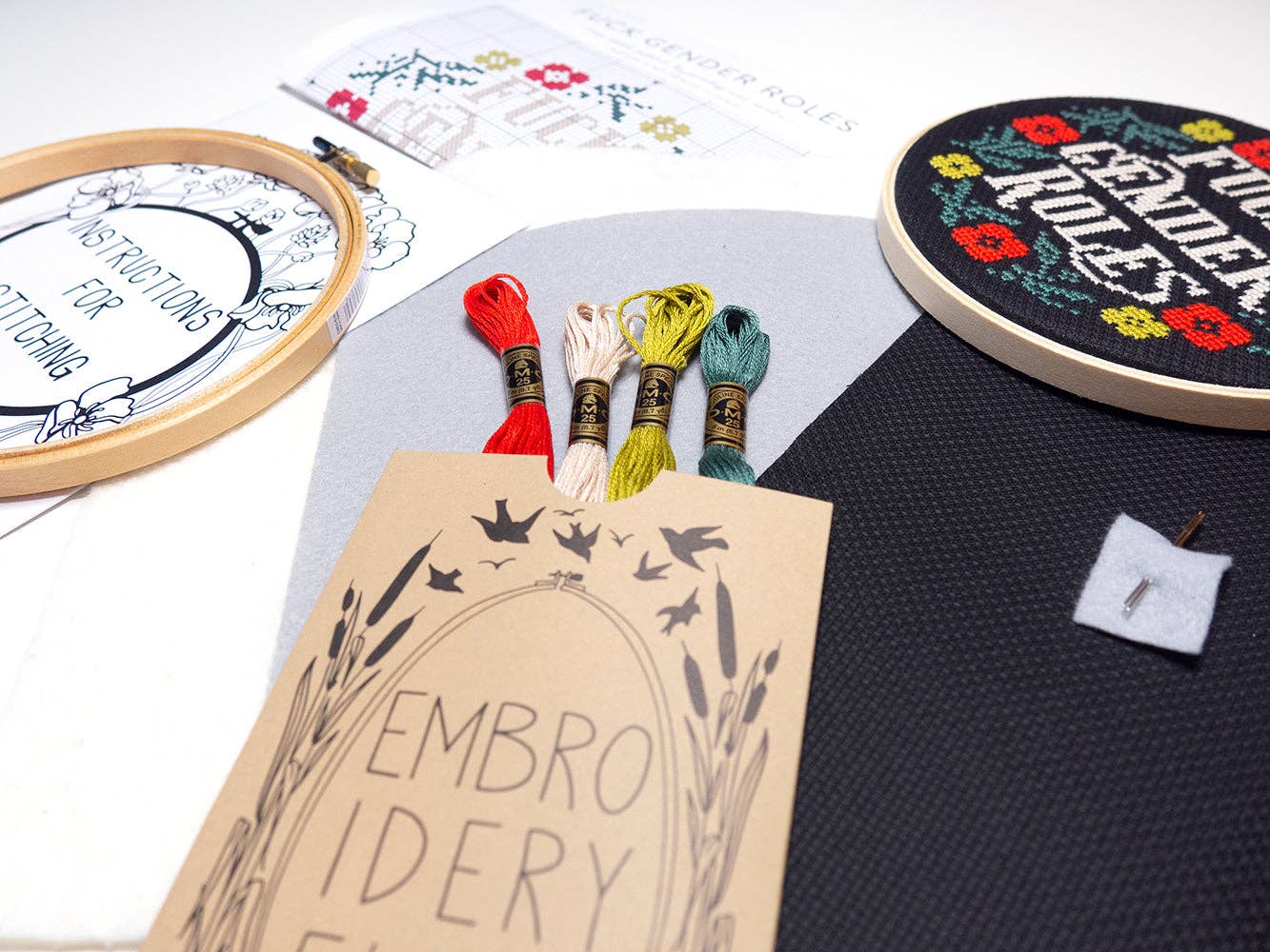 The Fuck Gender Roles Cross Stitch Kit is beginner-friendly and includes a hoop, colorful threads, printed instructions, needle, and a finished cross-stitch piece with “END GENDER ROLES” and floral designs.