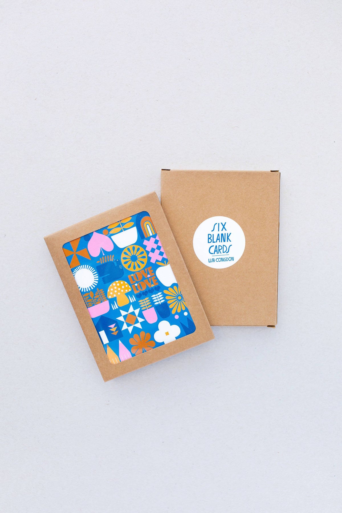 A brown envelope with the Love Love Mushroom Greeting Card, featuring colorful abstract geometric shapes, sits beside a closed brown box labeled SIX BLANK CARD LLR COINSHOP on a light gray surface.
