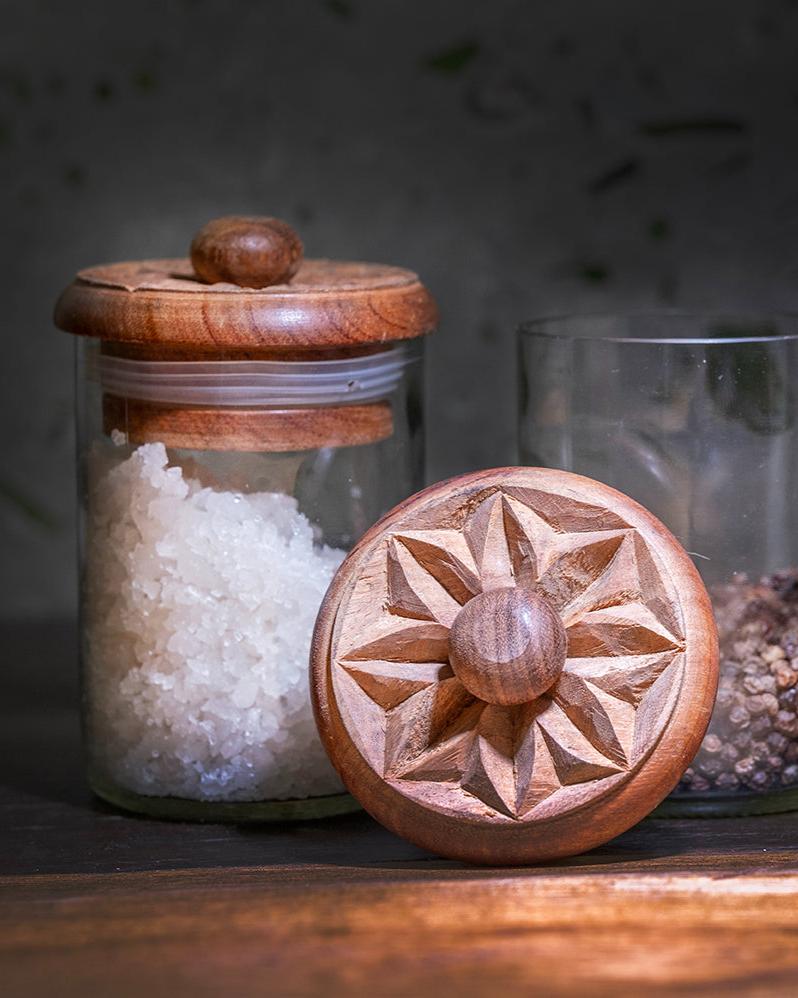 The Karafuu Upcycled Glass Jar | X Large, crafted by a Zanzibar artisan and filled with coarse salt, stands beside a peppercorn jar. Its wooden lid features an intricately carved geometric star pattern, showcasing skilled craftsmanship.