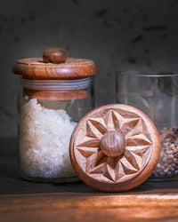 The Karafuu Upcycled Glass Jar | X Large, crafted by a Zanzibar artisan and filled with coarse salt, stands beside a peppercorn jar. Its wooden lid features an intricately carved geometric star pattern, showcasing skilled craftsmanship.