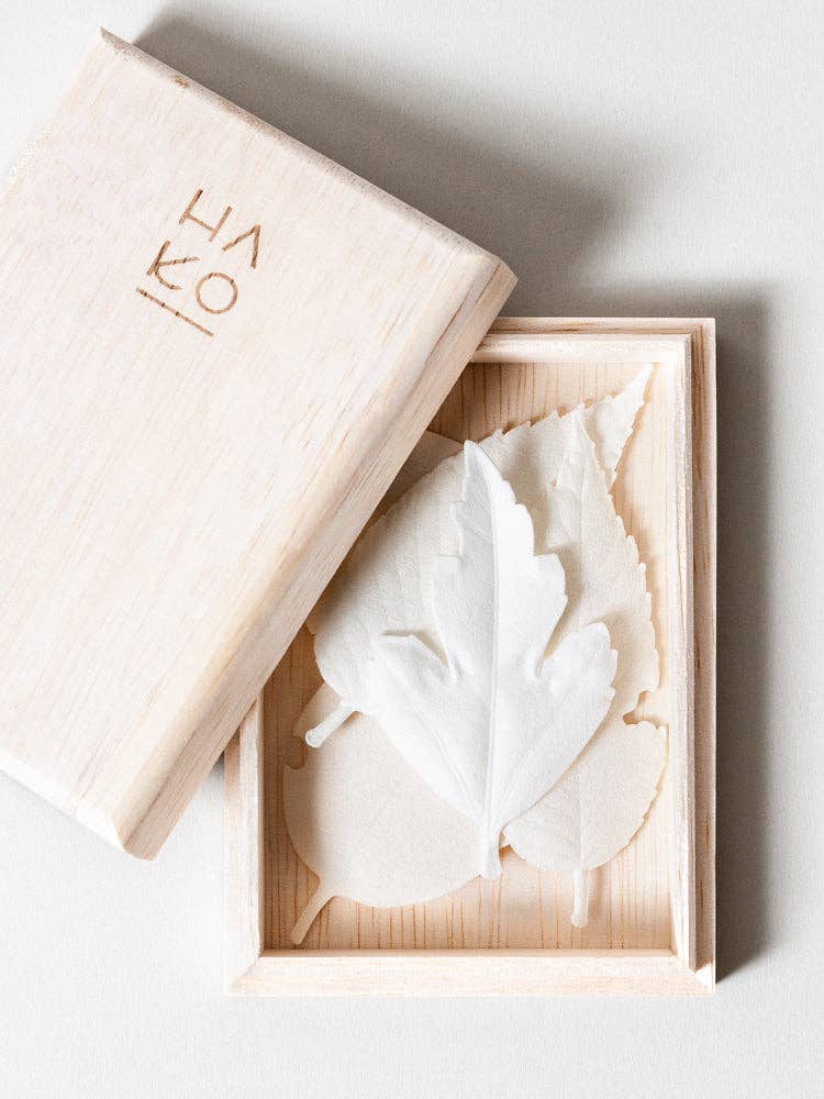 A light wooden box with a partially open lid reveals delicate, white HA KO Paper Incense inside. The elegant incense gift set features leaf-shaped papers and a lid stamped with Japanese characters, arranged neatly on a white surface.