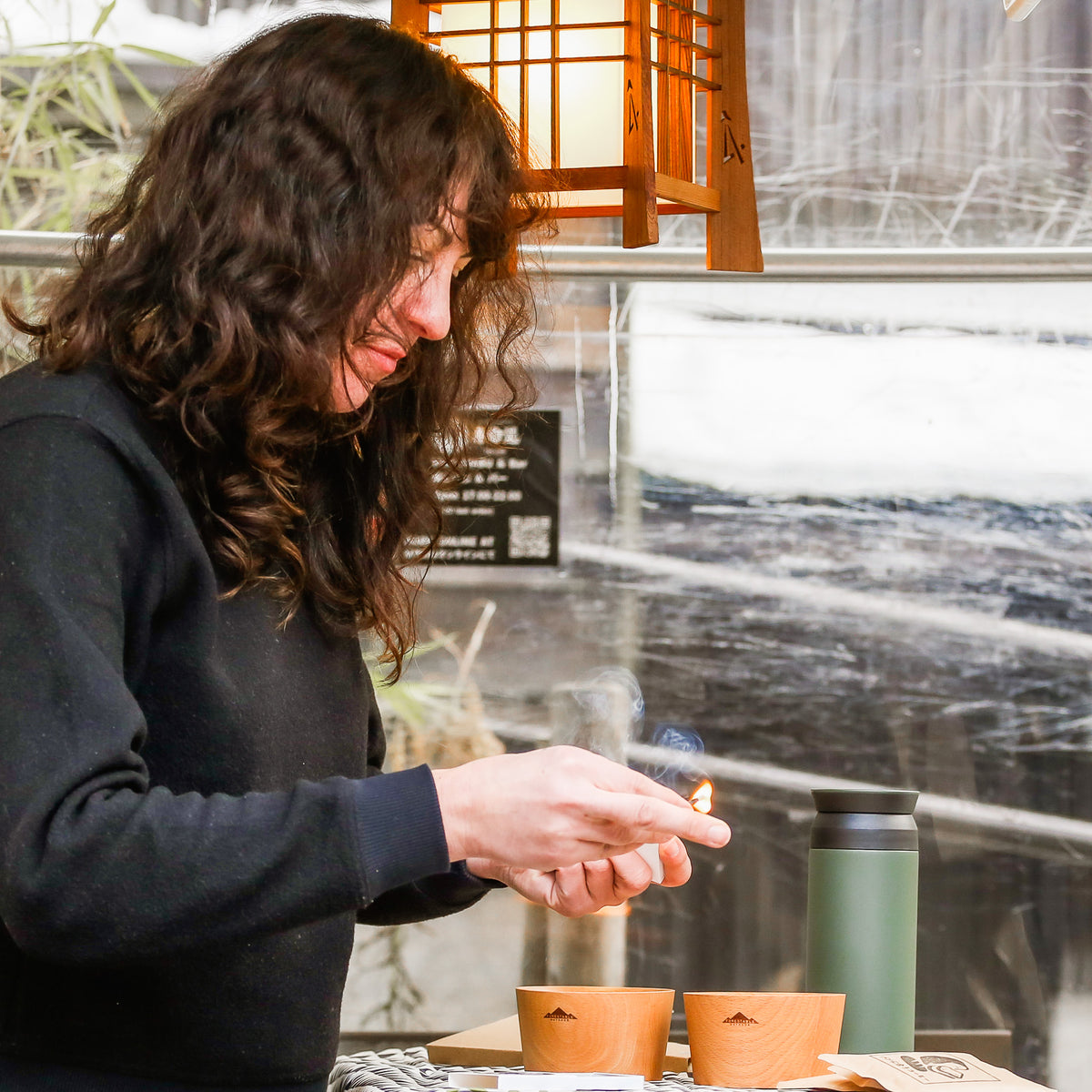 A person with long curly hair lights Hibi Japanese Incense Matches beside wooden cups and a thermos on a table under a hanging wooden lantern.