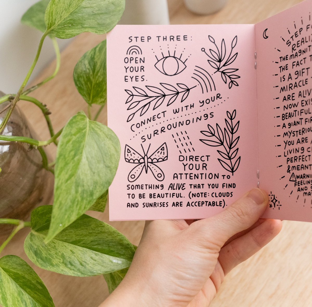 A hand holds open the Little Happiness Zine, a handmade booklet with handwritten text and doodles about mindfulness—featuring drawings of an eye, sun, butterfly, and leafy patterns—beside a green plant on a wooden surface.
