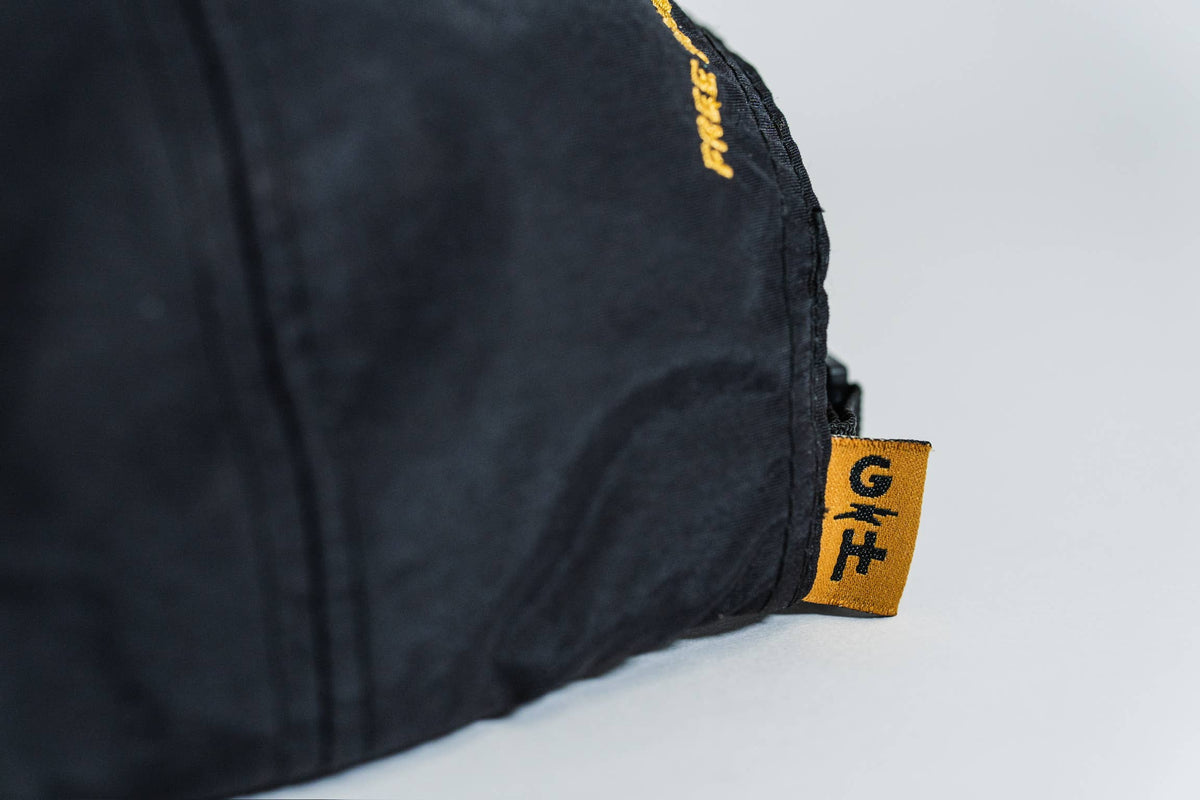 Close-up of the black Fearless 5-Panel Hat with a small yellow tag featuring "G H" and a line below. Some yellow "Gritty Habits" text is partially visible on the fabric in the background.