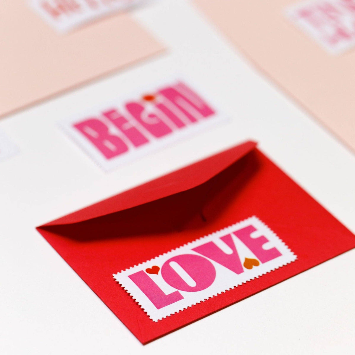 A red envelope with a white “LOVE” label and heart accents rests on a white surface beside Poster Stamp Series 5 | Words to Live By and pastel cards inspired by Lisa Congdon’s style.