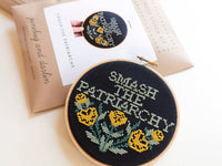 The Smash the Patriarchy Cross Stitch Kit is beginner-friendly, featuring yellow flowers and green leaves on black fabric with "SMASH THE PATRIARCHY" in bold. The packaging shows a photo of the finished design in the background.