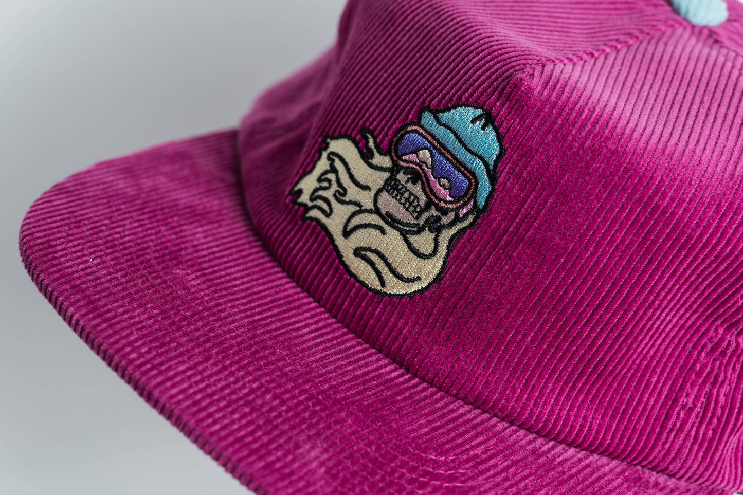 The Shred Til She's Dead Corduroy 5-Panel Hat showcases a magenta corduroy design with an embroidered grinning skull in blue goggles and helmet, flowing beige hair, and a moisture-wicking sweatband on the front panel.