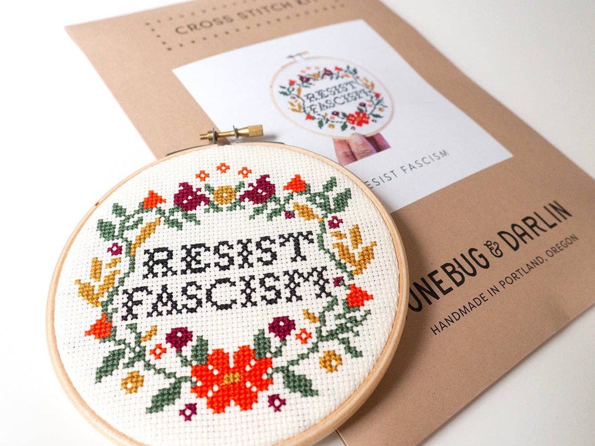 The beginner-friendly Resist Fascism Cross Stitch Kit features a finished hoop with "RESIST FASCISM" surrounded by colorful flowers, plus a pattern and packaging marked JUNebug & Darlin, handmade in Portland, Oregon.