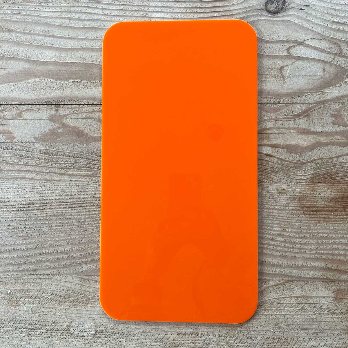 A bright orange rectangular object with rounded corners, likely The Mother | Home Base tote insert by Buena Goods, rests on a light wood-grain surface. Its glossy finish reflects part of a person holding a phone at the center.