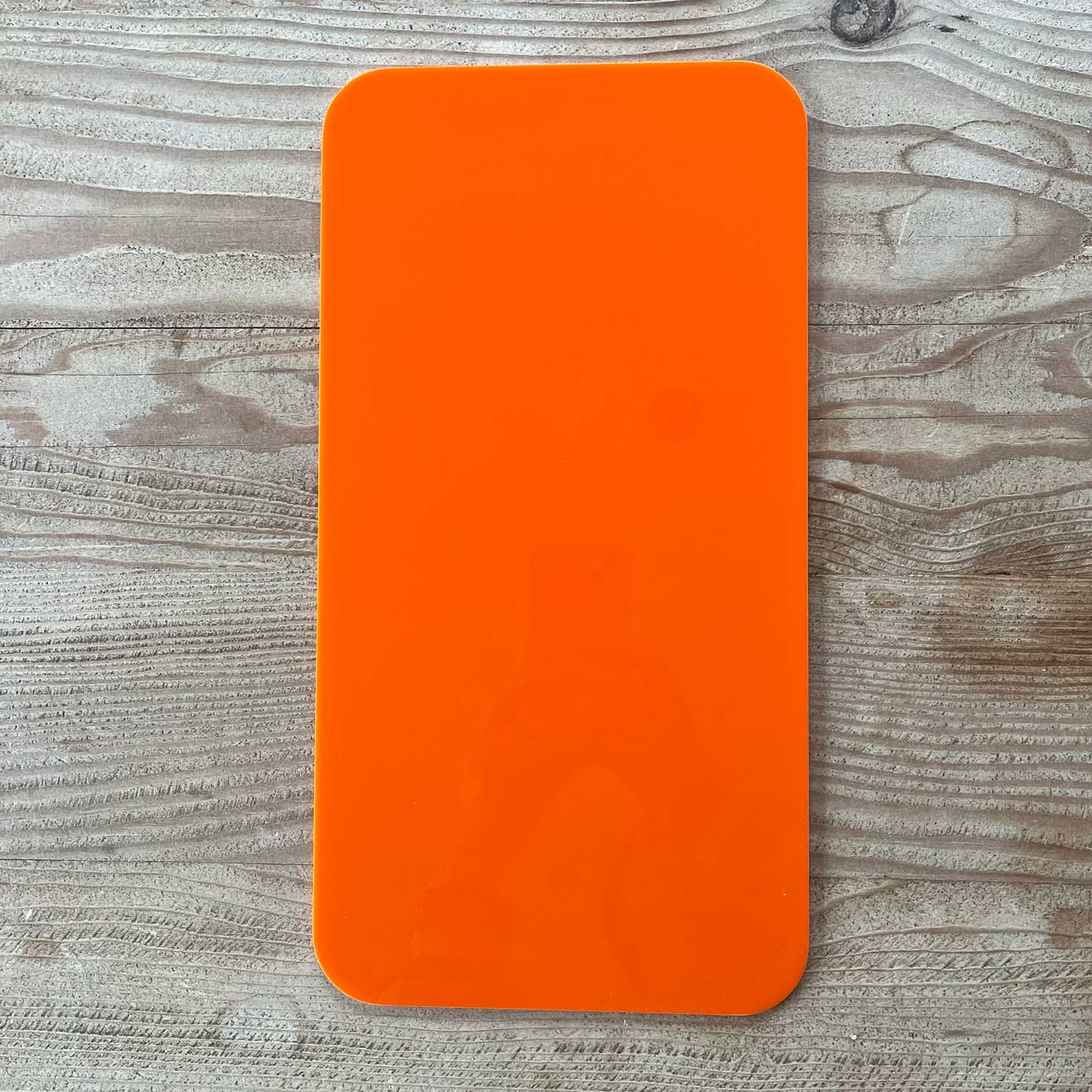 A bright orange rectangular object with rounded corners, likely The Mother | Home Base tote insert by Buena Goods, rests on a light wood-grain surface. Its glossy finish reflects part of a person holding a phone at the center.