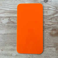 A bright orange rectangular object with rounded corners, likely The Mother | Home Base tote insert by Buena Goods, rests on a light wood-grain surface. Its glossy finish reflects part of a person holding a phone at the center.