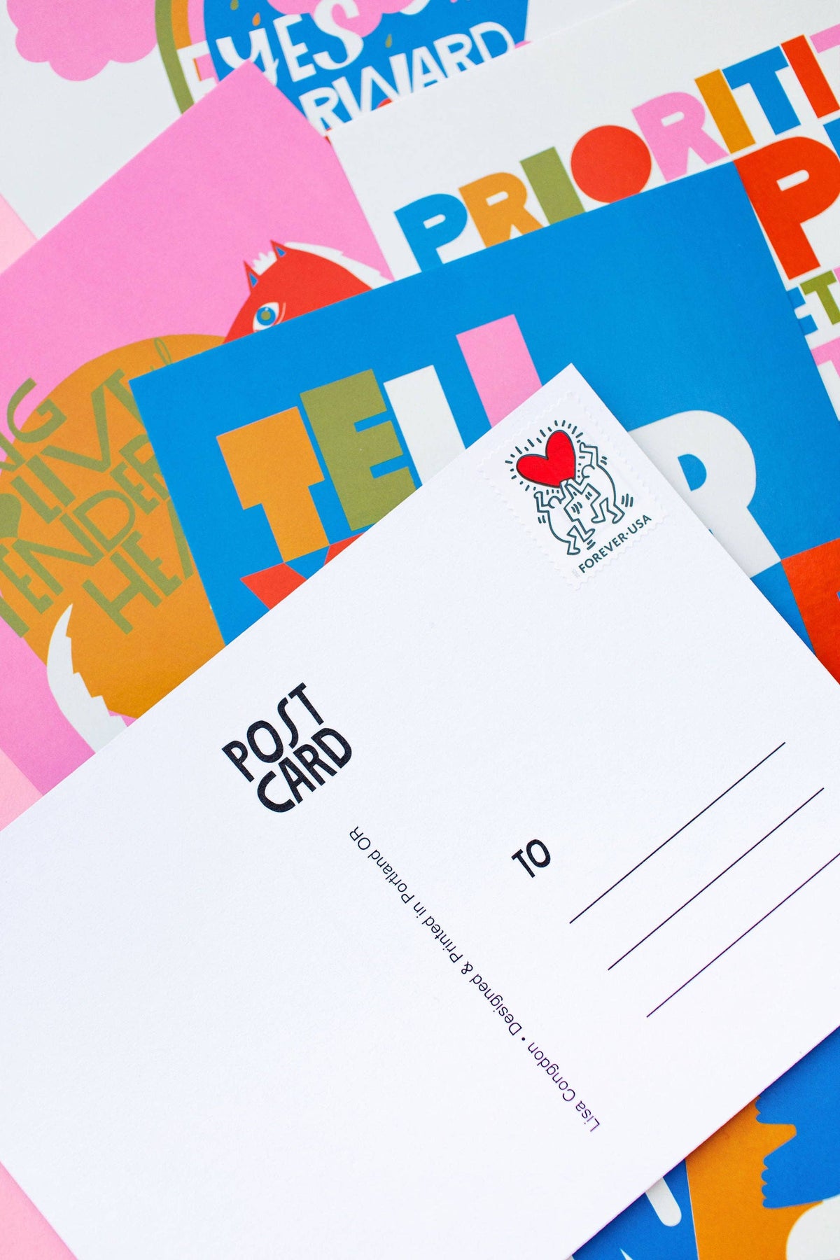 The XL Postcard Set features blank postcards with address lines and a small heart illustration, resting on vibrant posters with bold text—perfect for sending uplifting messages. All postcards are printed in Portland.