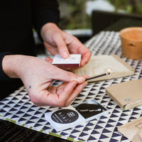 A person holds a lit match over a patterned surface, with Hibi Japanese Incense Matches, travel incense sticks, envelopes, and a small terracotta pot visible nearby.