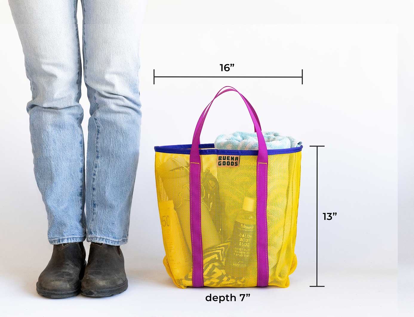 A person stands next to The Go Getter | Deep Blue, a durable mesh tote with purple handles. This reusable grocery bag measures 16" W x 13" H x 7" D and is partially filled with items.
