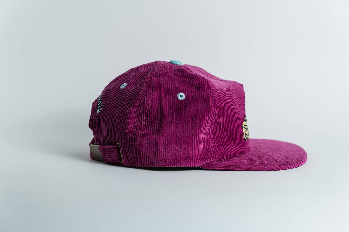 The Shred Til She's Dead Corduroy 5-Panel Hat in magenta, with blue eyelets, a light adjustable strap, and moisture-wicking sweatband, is shown on a plain white background.
