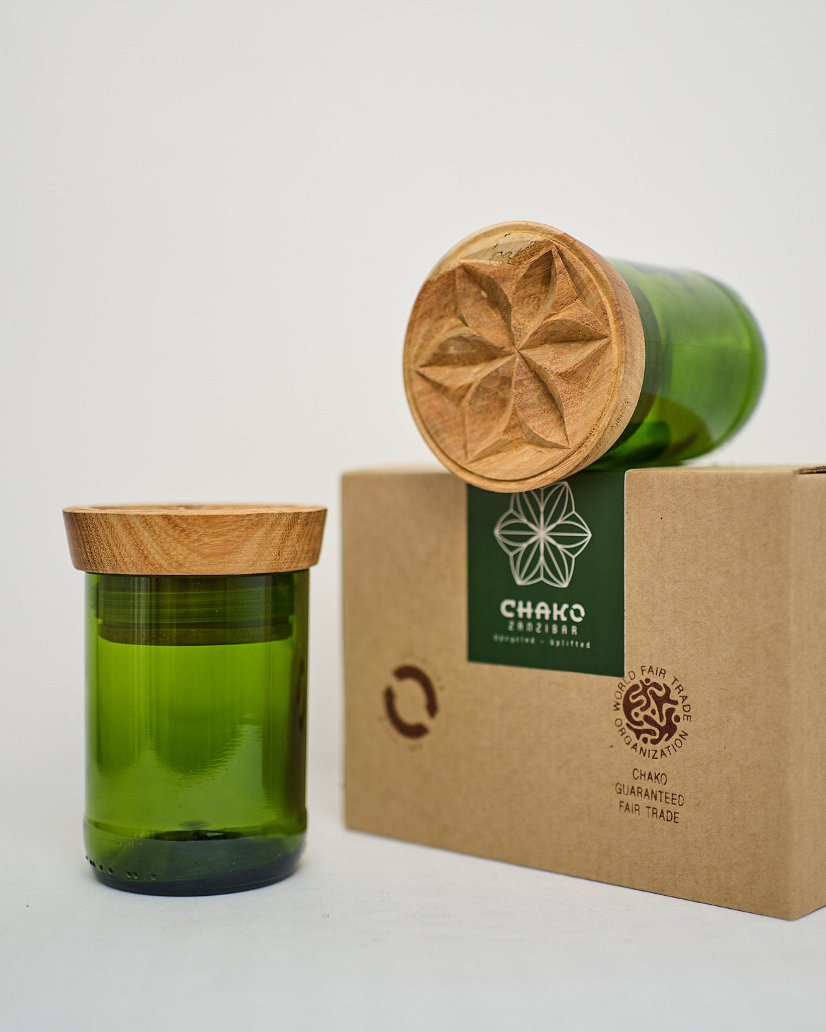 The Hiliki Upcycled Glass Jars | Large Set features two upcycled glass jars with carved wooden lids—one upright, one lying down—displayed on a brown box with CHAKO branding, a flower logo, and fair trade and recycling symbols.