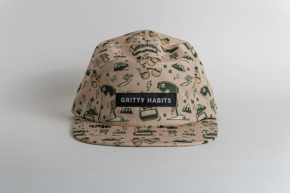 The First Chair 5-Panel Hat is beige, water-resistant, and features black and green cartoon-style illustrations with a black patch reading “GRITTY HABITS” in white. It’s shown against a plain white background.