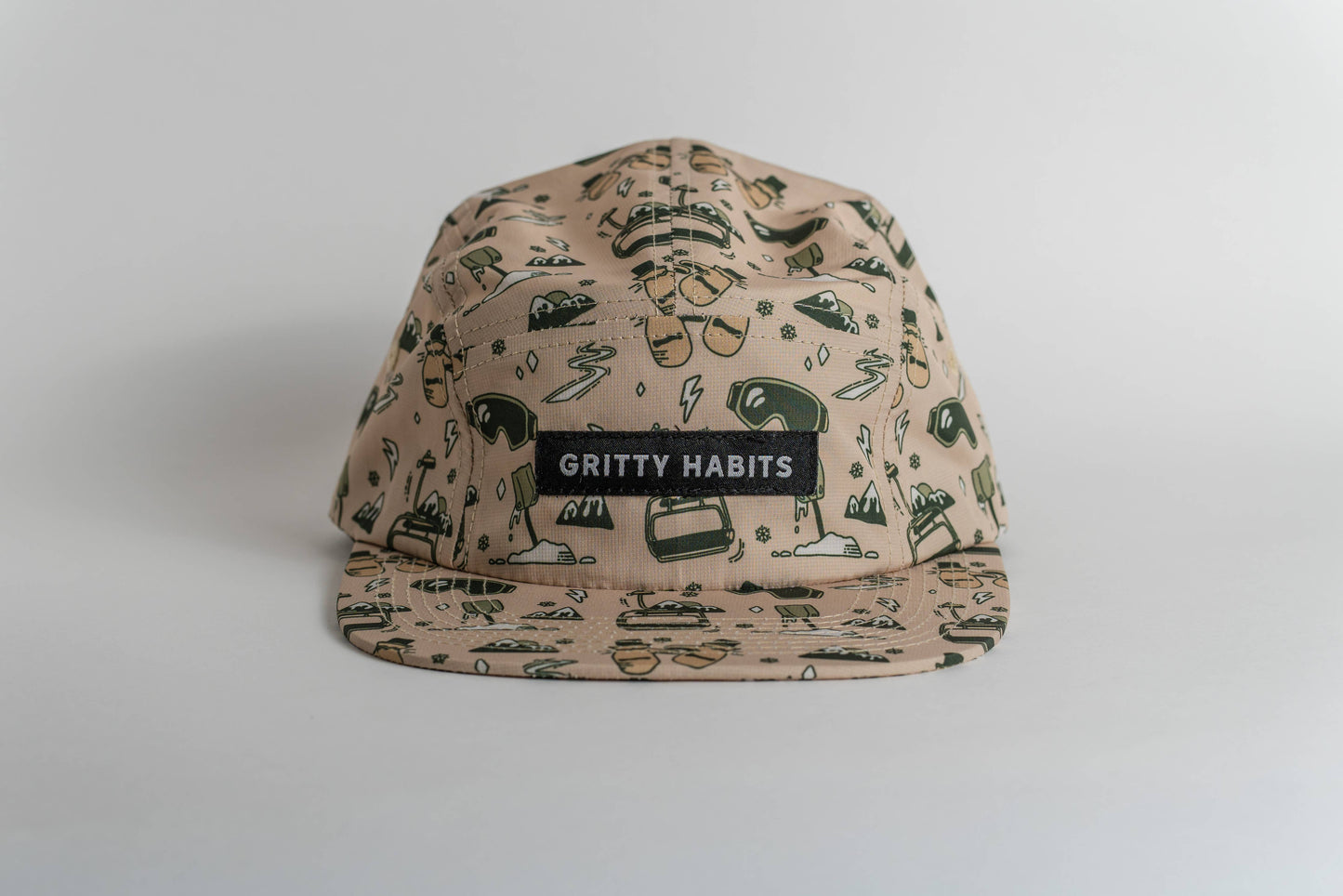 The First Chair 5-Panel Hat is beige, water-resistant, and features black and green cartoon-style illustrations with a black patch reading “GRITTY HABITS” in white. It’s shown against a plain white background.